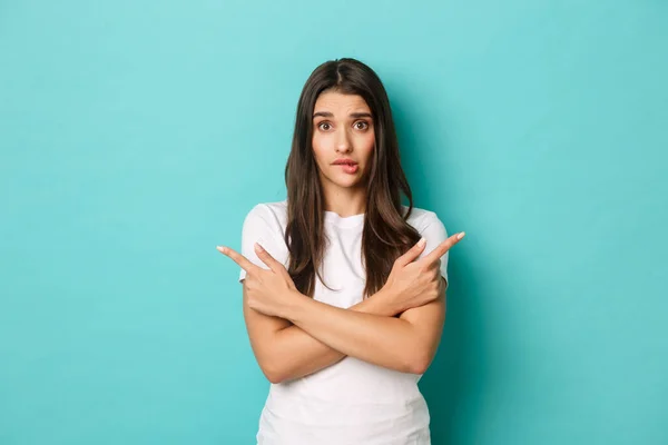 Portrait of cute and silly girl with long dark hairstyle, pointing sideways at copy space, asking for advice with choice, standing over blue background — Stock Photo, Image