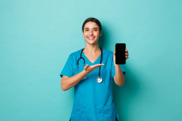 Covid-19, pandemia y concepto de medicina. Hermosa doctora, sonriendo y demostrando aplicación de teléfono móvil, mostrando aplicación de chequeo en línea, de pie sobre fondo azul — Foto de Stock