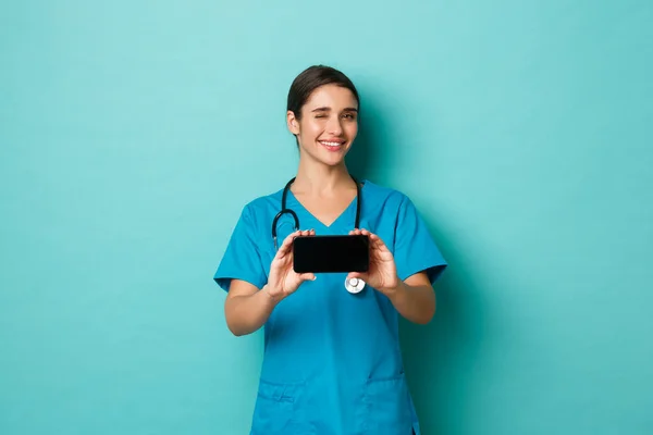 Coronavirus, pandemia y distanciamiento social. Imagen de una doctora sonriente en matorrales, guiñando un ojo y mostrando la pantalla del teléfono inteligente horizontalmente, de pie sobre fondo azul — Foto de Stock