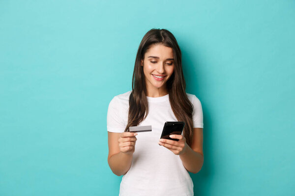Portrait of young brunette woman in casual white t-shirt, shopping online, holding credit card and mobile phone, standing over blue background