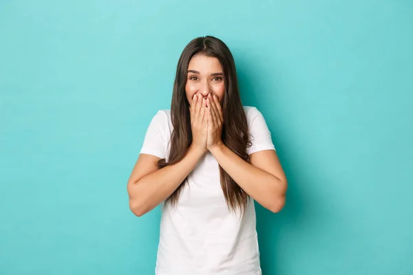 Portrait of surprised and excited brunette girl, looking amazed, cover mouth with hands, standing over blue background — Stock Photo, Image