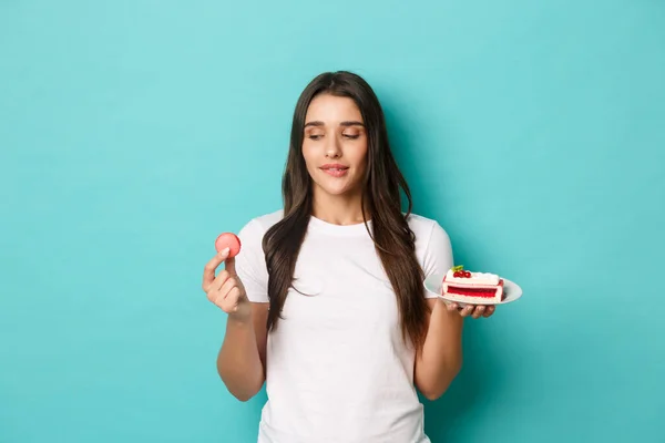 Imagen de una joven delgada en camiseta blanca, tentada a comer macarrones, sosteniendo pastel, de pie sobre fondo azul — Foto de Stock