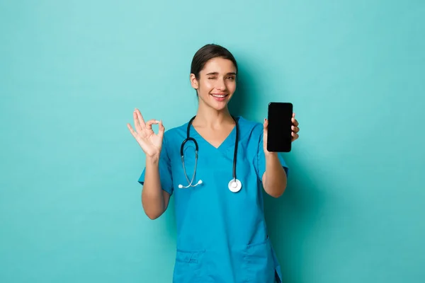 Covid-19, pandemia y concepto de medicina. Imagen de la bella doctora sonriente, vistiendo uniformes, mostrando la pantalla del teléfono móvil y el signo de aprobación, recomienda la aplicación, fondo azul — Foto de Stock