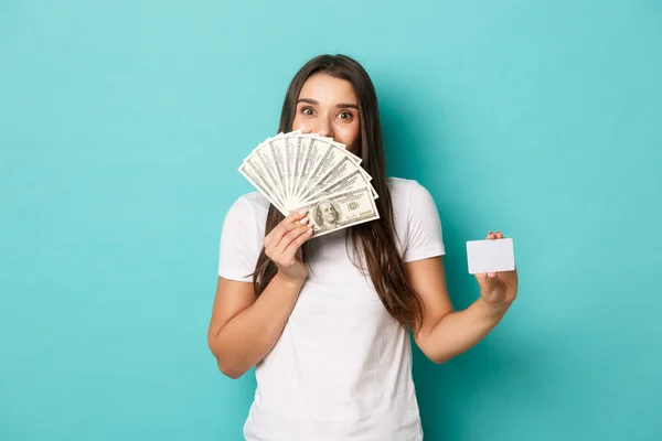 Изображение возбужденной улыбающейся женщины, чувствующей себя счастливой, показывающей кредитную карту и деньги, стоящей на синем фоне — стоковое фото
