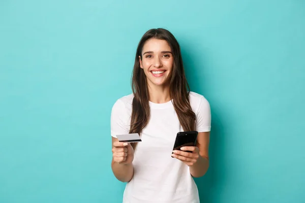 Porträtt av ung brunett kvinna i casual vit t-shirt, handla online, hålla kreditkort och mobiltelefon, stående över blå bakgrund — Stockfoto