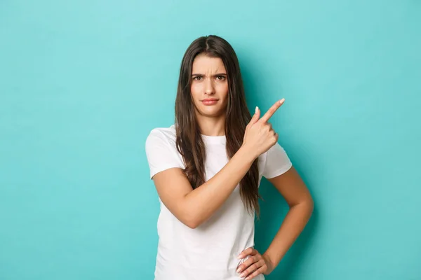 Image of confused and disappointed pretty girl in white t-shirt, frowning and pointing fingers at upper right corner, standing over blue background — Stock Photo, Image