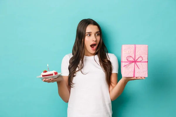 Young excited woman celebrating her birthday, holding bday cake and gift, standing amazed over blue background — Stock Photo, Image