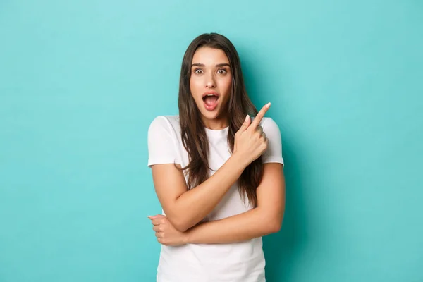 Cheerful brunette girl in white t-shirt, smiling excited and pointing fingers at upper right corner, showing amazing logo, standing over blue background — Stock Photo, Image