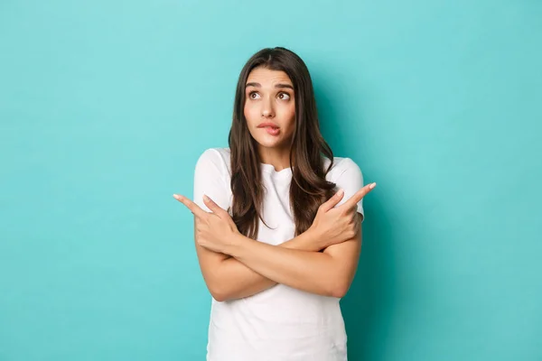 Portret van besluiteloos dom meisje in wit t-shirt, beslissingen nemen, vingers zijwaarts wijzen en naar links kijken, over een blauwe achtergrond staan — Stockfoto