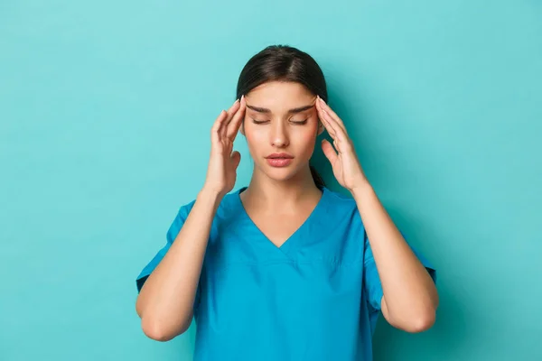 Coronavírus, distanciamento social e conceito de saúde. Close-up de mulher médico em esfrega se sentindo exausto, tendo uma dor de cabeça, de pé sobre fundo azul — Fotografia de Stock
