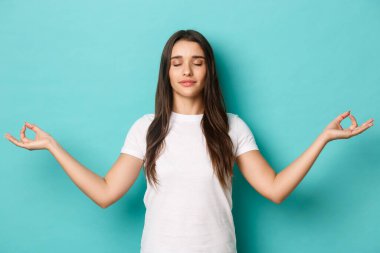 Beyaz tişörtlü barışçıl bir kız görüntüsü, gözlerini kapat ve ellerini yana aç, meditasyon yap, mavi arka planda yoga yap.