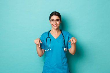 Coronavirus, salgın hastalık ve sosyalleşme konsepti. Önlüklü çekici bir kadın doktor resmi, parmaklarını aşağıya doğru gösteriyor ve gülümsüyor, logoyu gösteriyor, mavi arka planda duruyor.