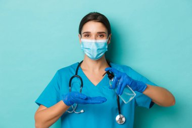 Covid-19 ve karantina kavramı. Tıbbi maskeli, eldivenli ve önlüklü, el dezenfektanı kullanan, mavi arka planda duran güzel kadın doktorların yakın çekimleri.
