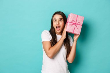 Tatil ve kutlama kavramı. Beyaz tişörtlü güzel bir kızın portresi hediye kutusunun içinde ne olduğunu tahmin etmeye çalışıyor, sallanıyor ve şaşırmış görünüyor, mavi arka planda duruyor.