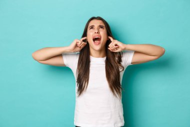 Öfkeli esmer kız bağırıyor ve hayal kırıklığına uğramış görünüyor, kulaklarını tıkamış rahatsız edici seslerden, komşulara bağırıyor, mavi arka planda dikiliyor.