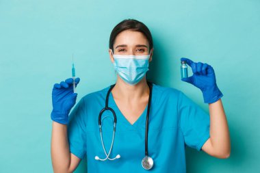 Covid-19 ve karantina kavramı. Tıbbi maskeli neşeli bayan doktor, lateks eldivenler ve önlükler, Coronavirus aşısı dolu şırınga gösteriyor, mavi arka planda duruyor.