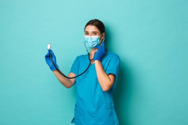 Covid-19, salgın hastalık ve ilaç konsepti. Tıbbi maskeli, eldivenli ve önlüklü güzel bir kadın doktor. Hastanın steteskopla kontrol etmesi ve kameraya gülümsemesi, mavi arka plan.