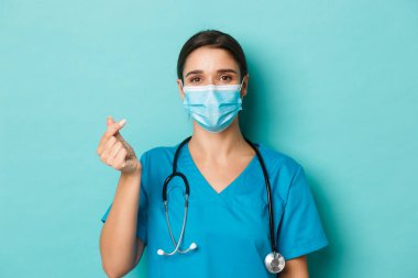 Covid-19 ve karantina kavramı. Ameliyat önlüğü ve tıbbi maske takmış neşeli, güzel bayan doktor, kalp işareti gösteriyor, mavi arka planda duruyor.