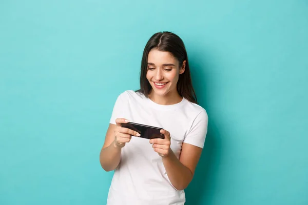 Bild av lycklig ung kvinna i vit t-shirt, spela mobiltelefon spel och leende, stående över blå bakgrund — Stockfoto