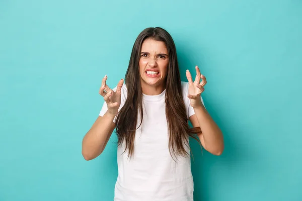 Immagine di giovane donna odiosa e arrabbiata in t-shirt bianca, pugni serrati e smorfie pazze, in piedi su sfondo blu — Foto Stock