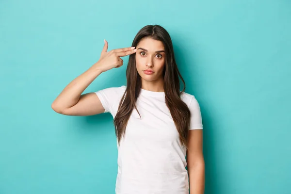 Image of annoyed and bothered female model, wearing white t-shirt, making finger gun sign near head, shooting herself from distress, standing over blue background — Stock Photo, Image