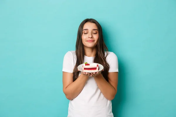 Imagen de tierna chica morena en camiseta blanca, mirando sabroso pedazo de pastel, de pie sobre fondo azul — Foto de Stock