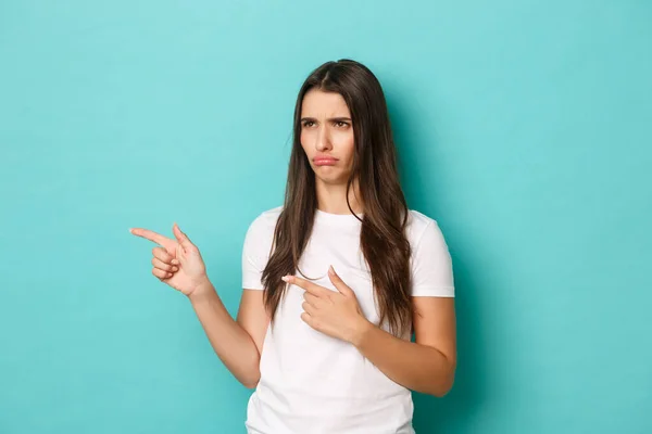 Portrait of disappointed pretty girl, complaining and sulking, looking and pointing fingers left at bad thing, standing over blue background — Stock Photo, Image