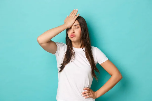 Young tired woman in white t-shirt, sighing and making facepalm to express annoyance, feeling fed up and tensed, standing over blue background — Stock Photo, Image