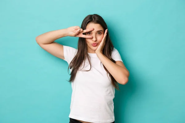 Imagem de menina glamour bobo em t-shirt branca, bebendo e mostrando sinal de paz sobre o olho, flertando sobre fundo azul — Fotografia de Stock