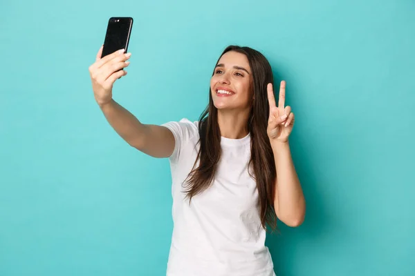 Porträtt av vacker lycklig kvinna i vit t-shirt, tar selfie med fred tecken, ler mot mobiltelefon, står över blå bakgrund — Stockfoto