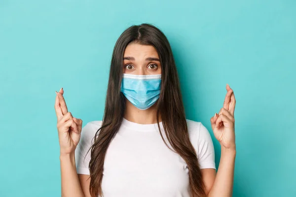 Conceito de coronavírus, saúde e distanciamento social. Close-up de menina preocupada em máscara médica, esperando e cruzando os dedos para a boa sorte, de pé sobre fundo azul — Fotografia de Stock