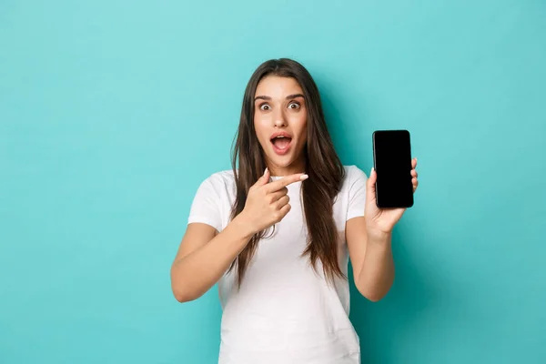 Retrato de mulher bonita espantada em t-shirt branca, apontando o dedo para a tela do telefone móvel, de pé animado sobre fundo azul — Fotografia de Stock