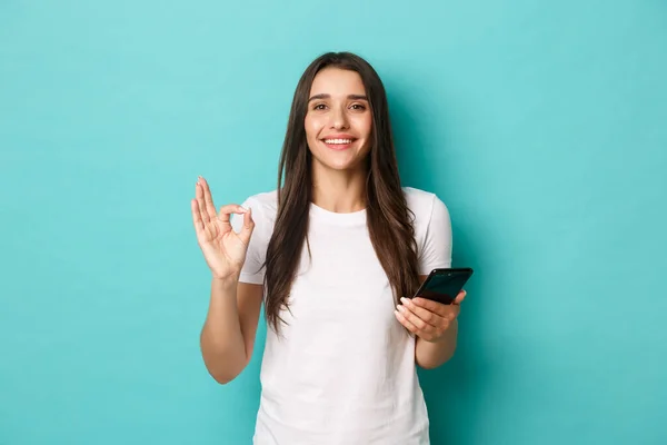 Vacker ung kvinna i vit t-shirt, håller mobiltelefon och visar okej tecken, rekommenderar något online, stående nöjd över blå bakgrund — Stockfoto