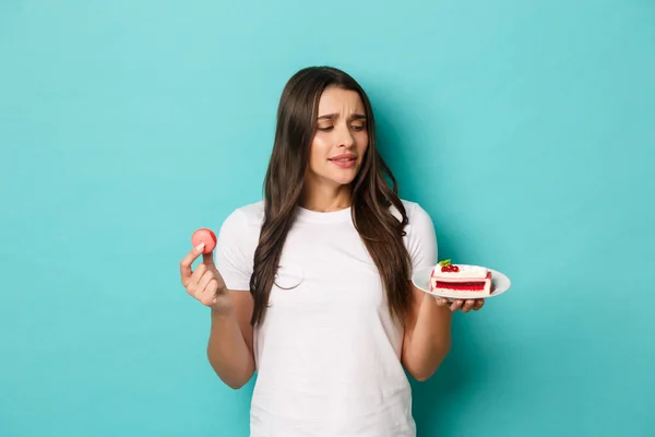 Imagen de una joven delgada en camiseta blanca, tentada a comer pastel, sosteniendo macarrones, de pie sobre fondo azul — Foto de Stock