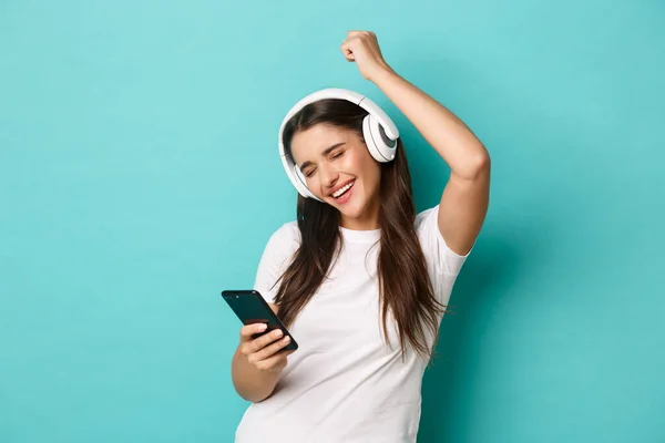 Portrait de jolie fille moderne en t-shirt blanc, écoutant de la musique dans des écouteurs sans fil, tenant le téléphone portable et dansant, debout sur fond bleu — Photo