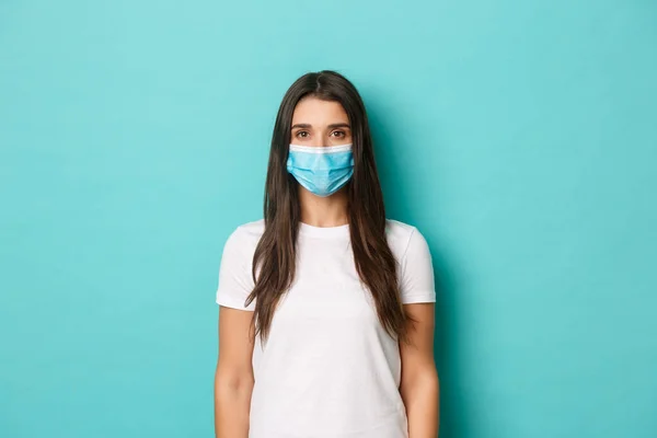 Begreppet pandemi, covid-19 och socialt avstånd. Ung beslutsam kvinna i vit t-shirt bär medicinsk mask, tittar på kameran allvarlig, står över blå bakgrund — Stockfoto