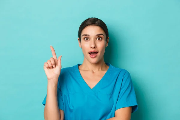 コロナウイルス、社会的距離と健康の概念。青い背景の上にスクラブに立って、指を上げて解決策を提案するアイデアを持っている興奮した女性医師のクローズアップ — ストック写真