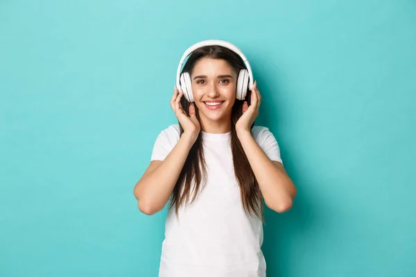 Retrato de una bonita joven en camiseta blanca, sonriendo y escuchando música en auriculares inalámbricos, de pie sobre fondo azul — Foto de Stock