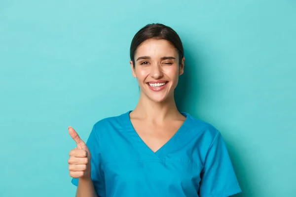 Coronavírus, distanciamento social e conceito de saúde. Close-up de médico feminino alegre, piscando e sorrindo, mostrando polegar-se em aprovação, de pé otimista sobre fundo azul — Fotografia de Stock