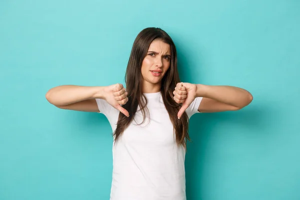 Imagen de una morena disgustada con una camiseta blanca básica, frunciendo el ceño y mirando a la crítica, mostrando los pulgares hacia abajo en algo muy malo, de pie sobre un fondo azul — Foto de Stock