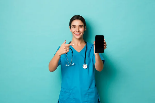 Covid-19, pandemia y concepto de medicina. Alegre médico sonriente en matorrales, señalando con el dedo el teléfono móvil, mostrando la aplicación, de pie sobre el fondo azul — Foto de Stock