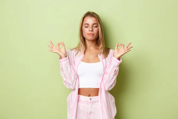 Retrato de mujer joven relajada con peinado corto rubio, ojos cerrados y cogidas de la mano en gesto zen, meditando, practicando yoga sobre fondo verde — Foto de Stock