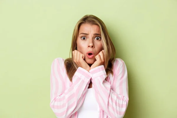 Close-up of shocked blond woman looking scared, standing ambushed with something scary over green background — Stock Photo, Image