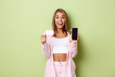 Pembe gömlekli ve kotlu şık sarışın bir kadın resmi. Akıllı ekranı ve kredi kartını gösteriyor. Çevrimiçi bir şey öneriyor.