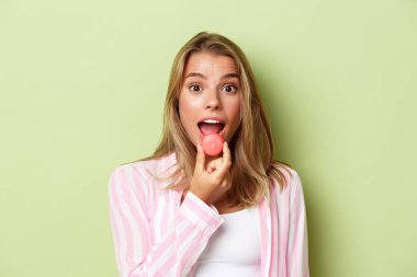 Pembe tişörtlü aptal sarışın kızın yakın plan fotoğrafı, badem kurabiyesi yiyor ve mutlu görünüyor, yeşil arka planda duruyor.