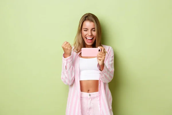 Retrato de menina vencedora feliz com cabelo loiro, olhando para a tela do smartphone e triunfando, de pé sobre fundo verde — Fotografia de Stock