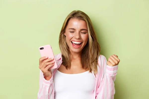 Крупный план счастливой блондинки достигает ежедневной цели в мобильном приложении, выигрывая что-то, держа смартфон и радуясь, стоя на зеленом фоне — стоковое фото