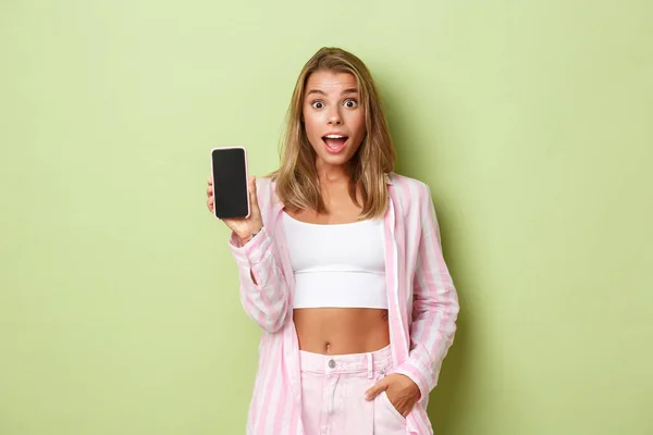 Imagem de modelo feminino atraente loiro em camisa rosa, mostrando tela do smartphone e olhando animado, de pé sobre fundo verde — Fotografia de Stock