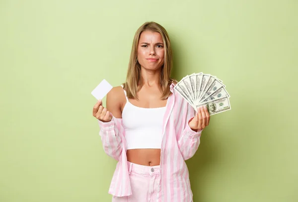 Immagine di donna bionda scettica che sembra confusa, accigliata perplessa, in possesso di carta di credito e contanti, in piedi su sfondo verde — Foto Stock