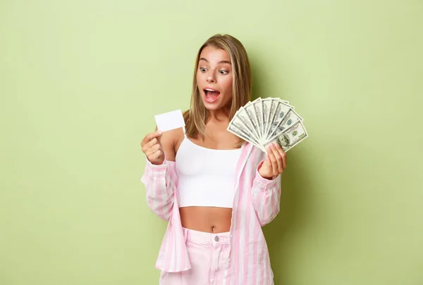 Ritratto di attraente donna ricca con i capelli biondi, con soldi in mano e carta di credito e sorridente stupito, in piedi su sfondo verde — Foto Stock